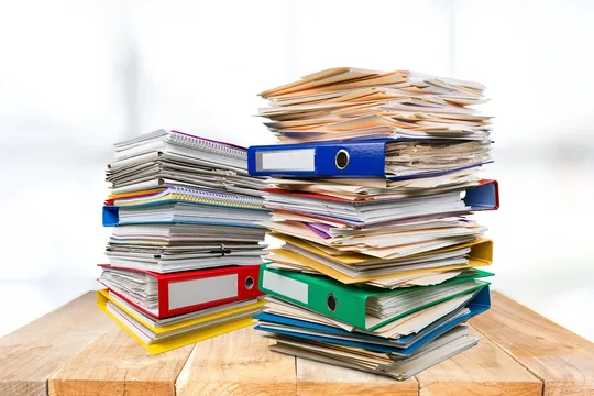 Two tall stacks of colourful binders and papers on a wooden desk
